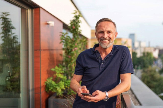 Ein Mann mit Bart steht auf einem Balkon und hält ein Handy in der Hand, während er die Aussicht genießt.