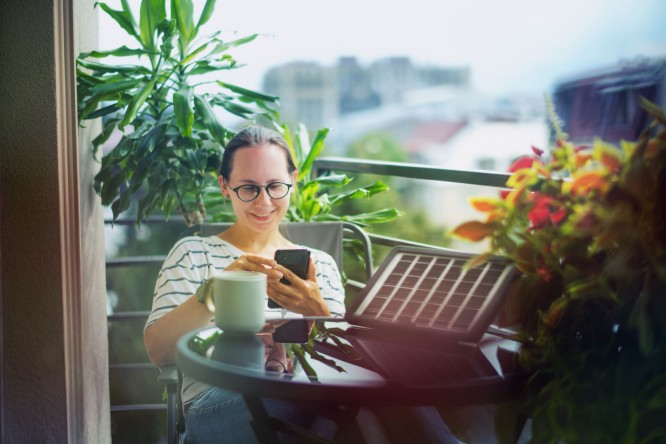 Eine Frau sitzt auf ihrem Balkon und nutzt Solarenergie über ein Solarpanel, während sie auf ihr Smartphone schaut. Einspeisevergütung Auri Sun